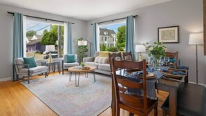 Een moderne woonkamer met een bank, stoelen, een salontafel en een eettafel voor vier personen. Grote ramen bieden uitzicht op de omgeving.