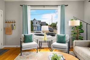 Ein Wohnzimmer mit zwei grauen Sesseln, einem Couchtisch, einer Lampe und Blick auf die Straße durch ein mit Vorhängen versehenes Fenster.