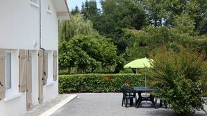 A building with a white facade and a gravel courtyard. There is a table with chairs and a green umbrella. - Chez Régine - Angoume (ANGOUME)