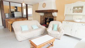 A living room with a sofa, armchair, and coffee table. There is a fireplace, kitchen area, and a dining table with chairs. - Chez Régine - Angoume (ANGOUME)
