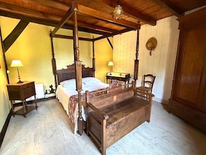 Un lit à baldaquin avec une tête de lit en bois, une table de chevet, une chaise et une armoire dans une pièce dotée de poutres apparentes et d’un plafond en pente.