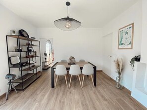 Un comedor con una mesa de madera y sillas blancas, una estantería metálica negra con diversos objetos, y una lámpara colgante.