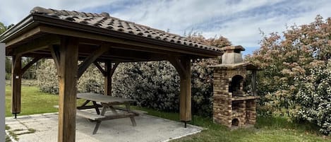 Un kiosque en bois avec une table de pique-nique, entouré d’un jardin agrémenté d’arbustes en fleurs.