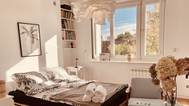 Ein modernes Schlafzimmer mit einem großen Bett, einem Nachttisch, einem Sessel, einem Bücherregal und einem Fenster mit Blick auf Bäume und Gebäude.