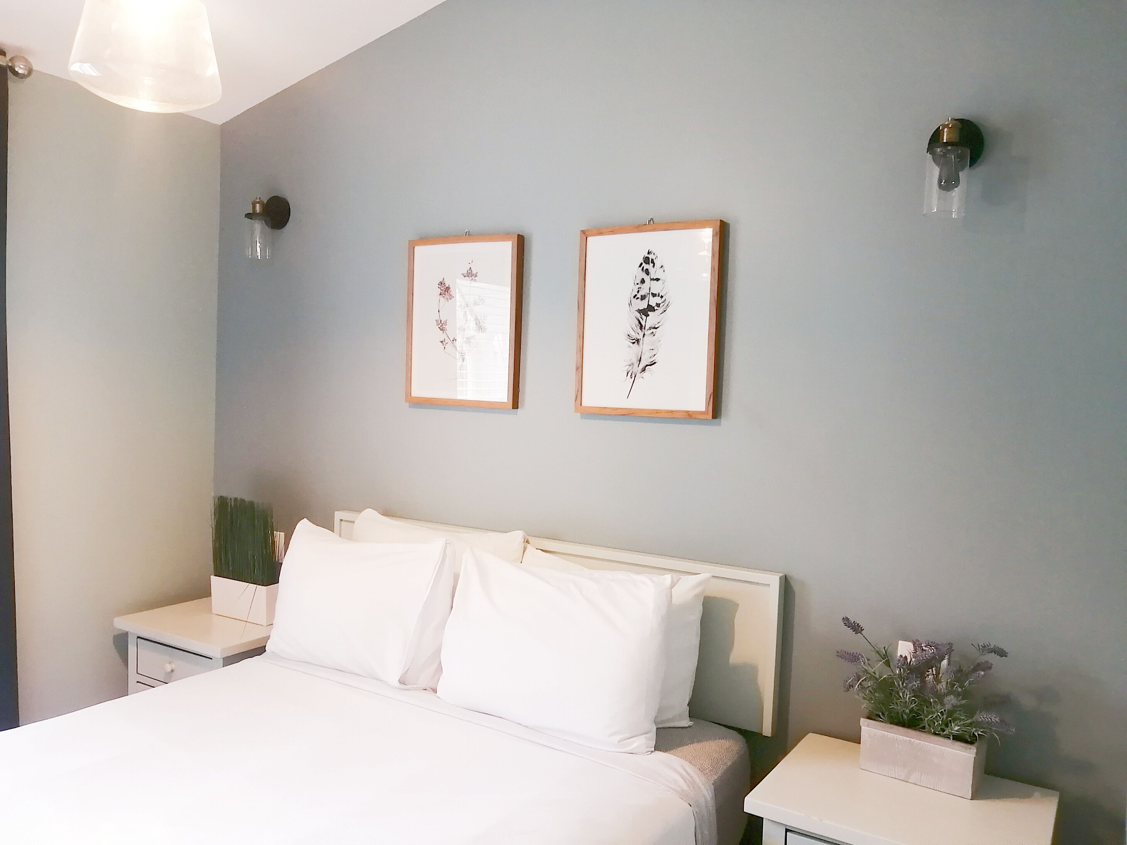 Un dormitorio con una cama, dos cuadros en la pared, una mesita de noche con una planta y una lámpara de pared.
