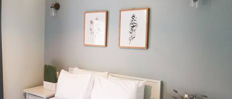 Un dormitorio con una cama, dos cuadros en la pared, una mesita de noche con una planta y una lámpara de pared.