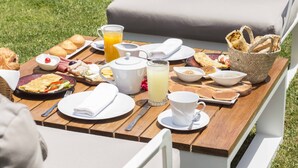 Una mesa de madera al aire libre con desayuno servido, incluyendo diversos alimentos, una jarra de jugo de naranja y una cesta de pan.