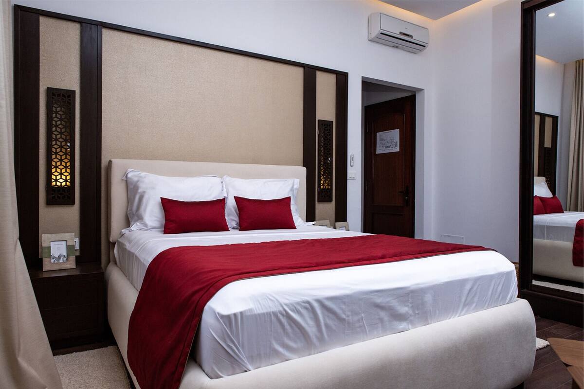 Una habitación de hotel con una cama grande, almohadas rojas, cabecera blanca y aire acondicionado instalado en la pared.