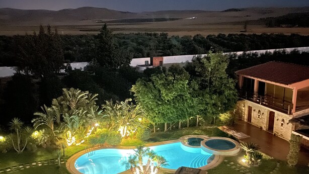 Vista nocturna de una zona de piscina con piscina circular, rodeada de árboles iluminados y un edificio de piedra.