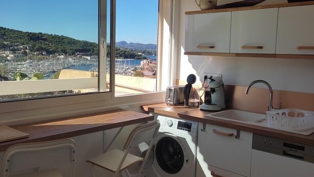 Een compacte keuken met een wasmachine, een spoelbak en een raam met uitzicht op een jachthaven en heuvels.