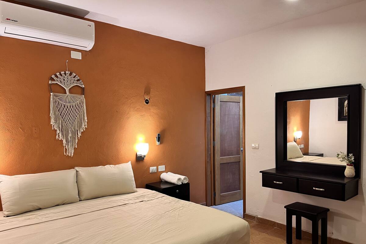 A hotel room with a bed, a wall-mounted macrame decoration, a wall-mounted light, a bedside telephone, a mirror, and a small table.