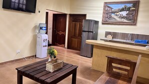 Een compacte keuken met een inbouwkoelkast, waterdispenser, een houten bankje en een betegelde vloer.