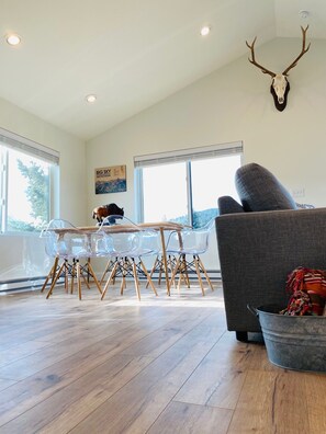 A modern living room with a wooden floor, a gray sofa, a clear glass table with wooden legs, and a mounted deer skull on the wall. - NEW Listing! NEW Build! Condo is 1-block walk to restaurants, shopping, concerts (Big Sky)