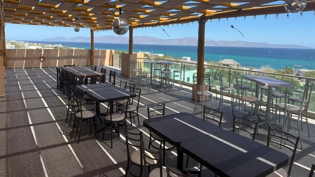 Eine Außenterrasse mit Tischen und Stühlen, einer Pergola und Blick auf das Meer und die Berge.