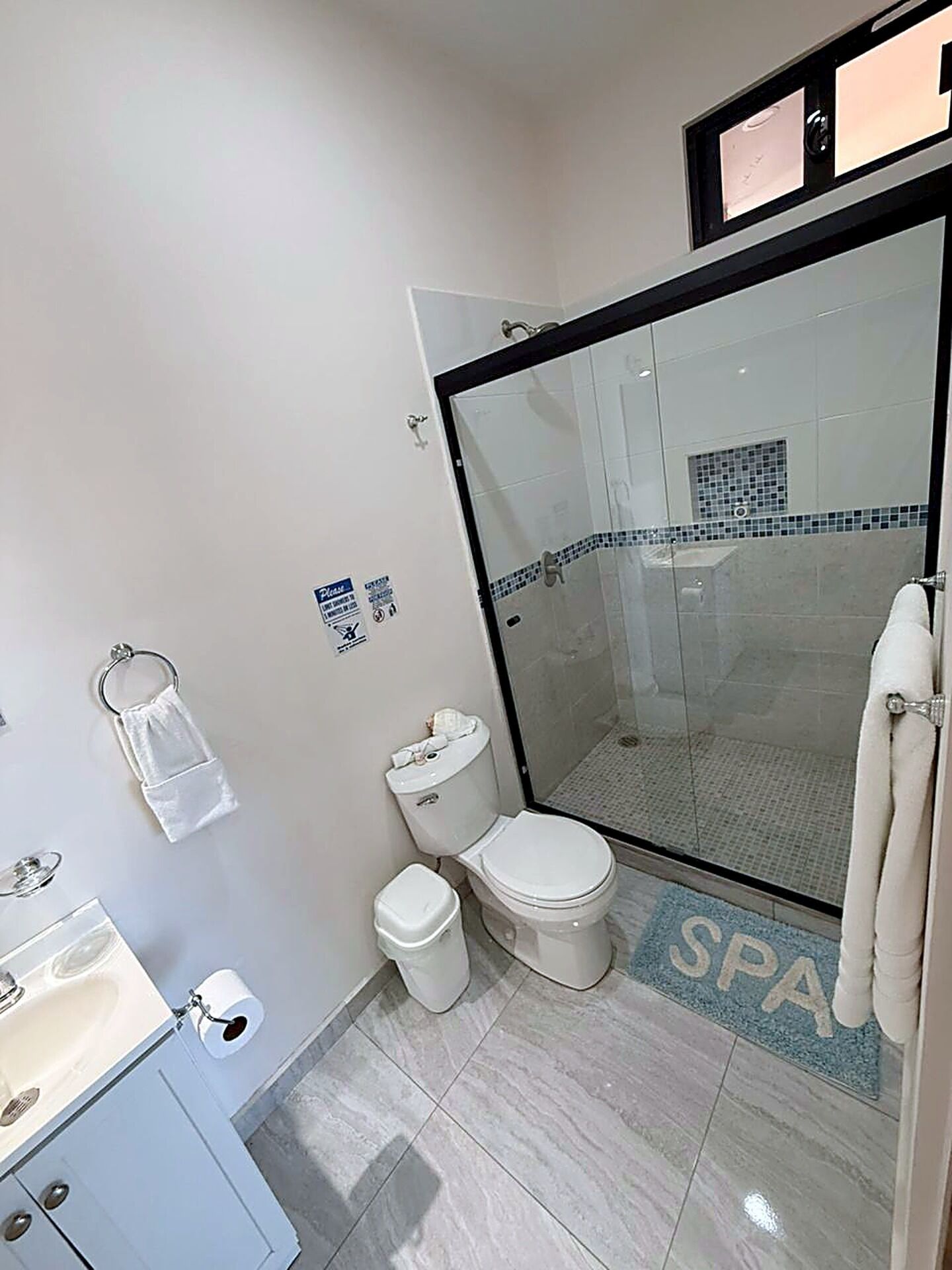 A bathroom with a toilet, a sink, a glass-enclosed shower, and a spa-themed floor mat.
