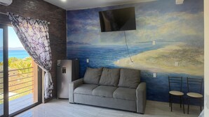 Ein Wohnzimmer mit grauem Sofa, einem flachbildfernseher an der Wand und einem großen Fenster mit Meerblick.