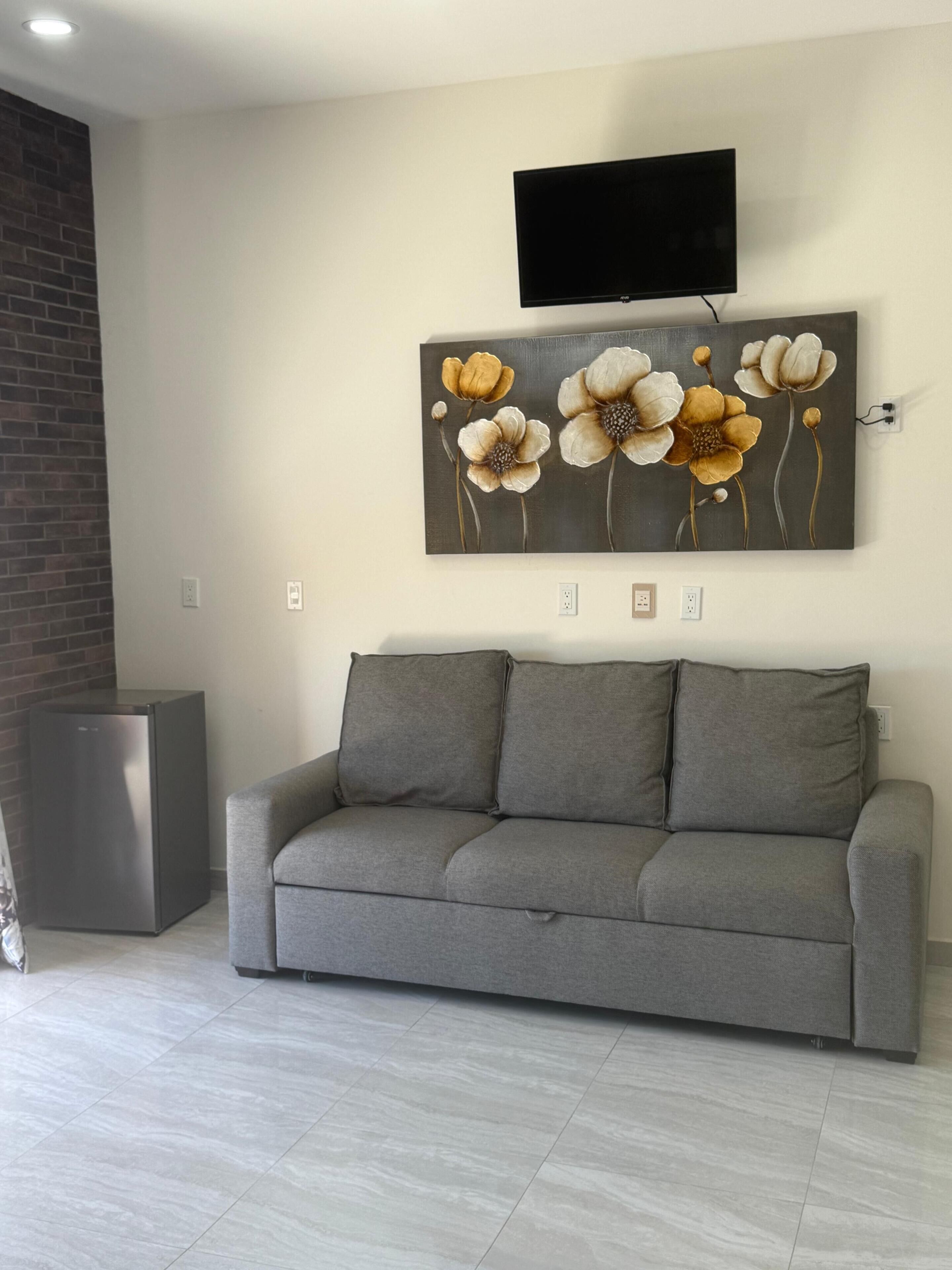 A living room with a gray sofa, a flat-screen TV mounted above a floral painting, and a brick accent wall.