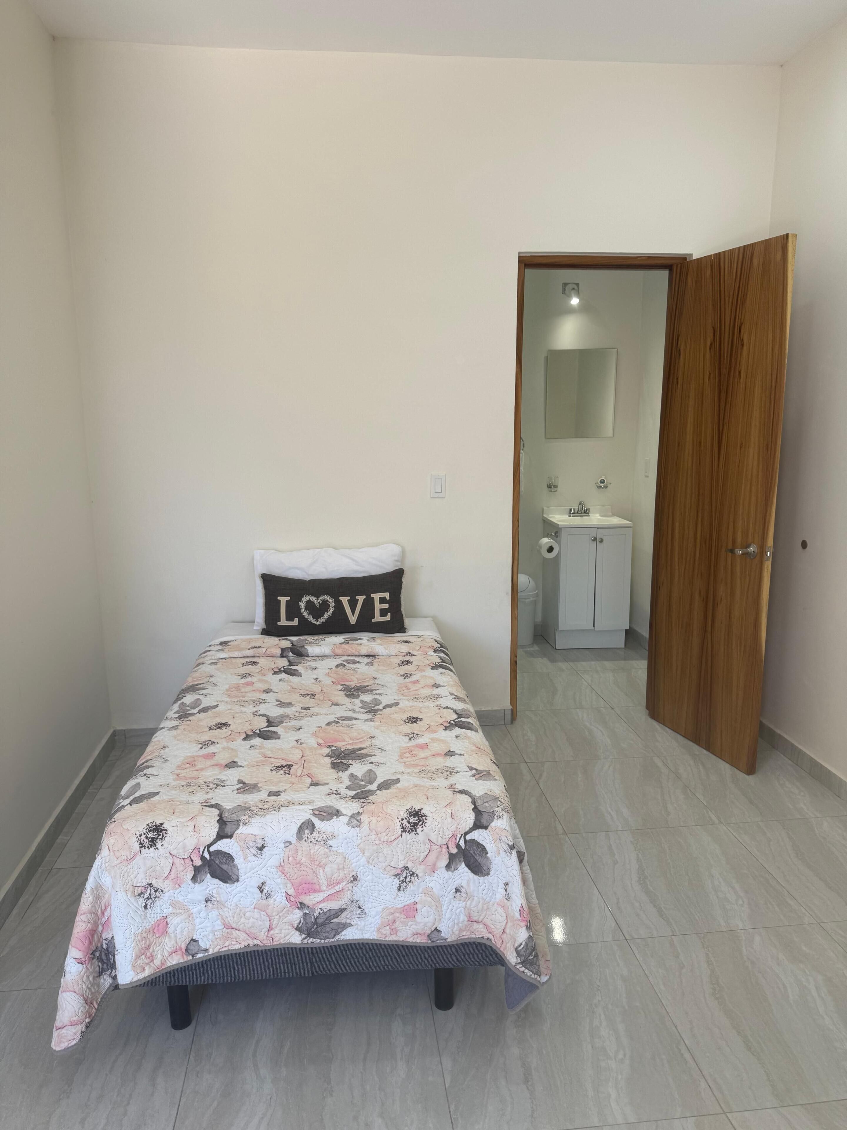 A bedroom with a bed, a pillow with 'LOVE' text, a wooden door, and a bathroom visible through an open door.