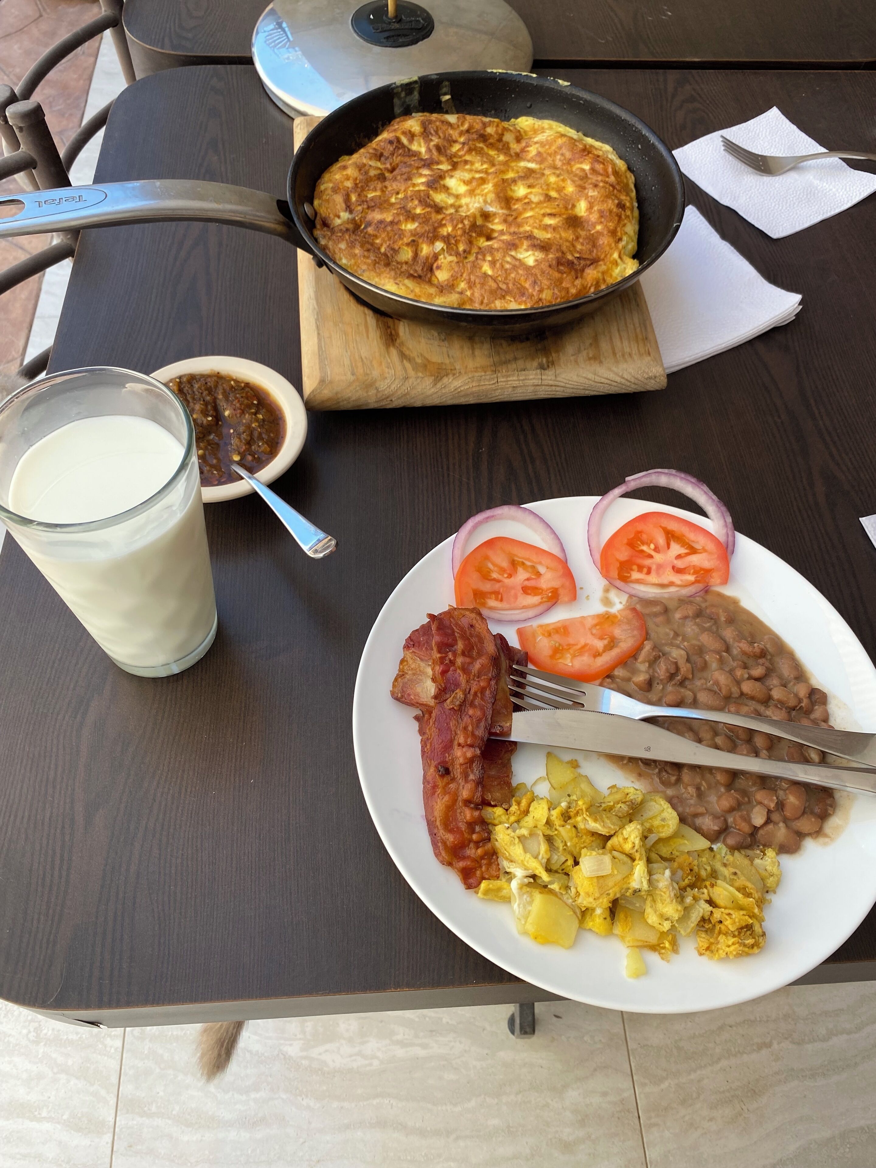 A breakfast plate with eggs, bacon, beans, tomatoes, and onions, a glass of milk, and a skillet with a dish on a wooden board.