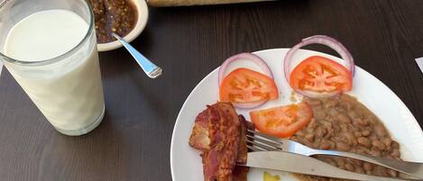 A breakfast plate with eggs, bacon, beans, tomatoes, and onions, a glass of milk, and a skillet with a dish on a wooden board.