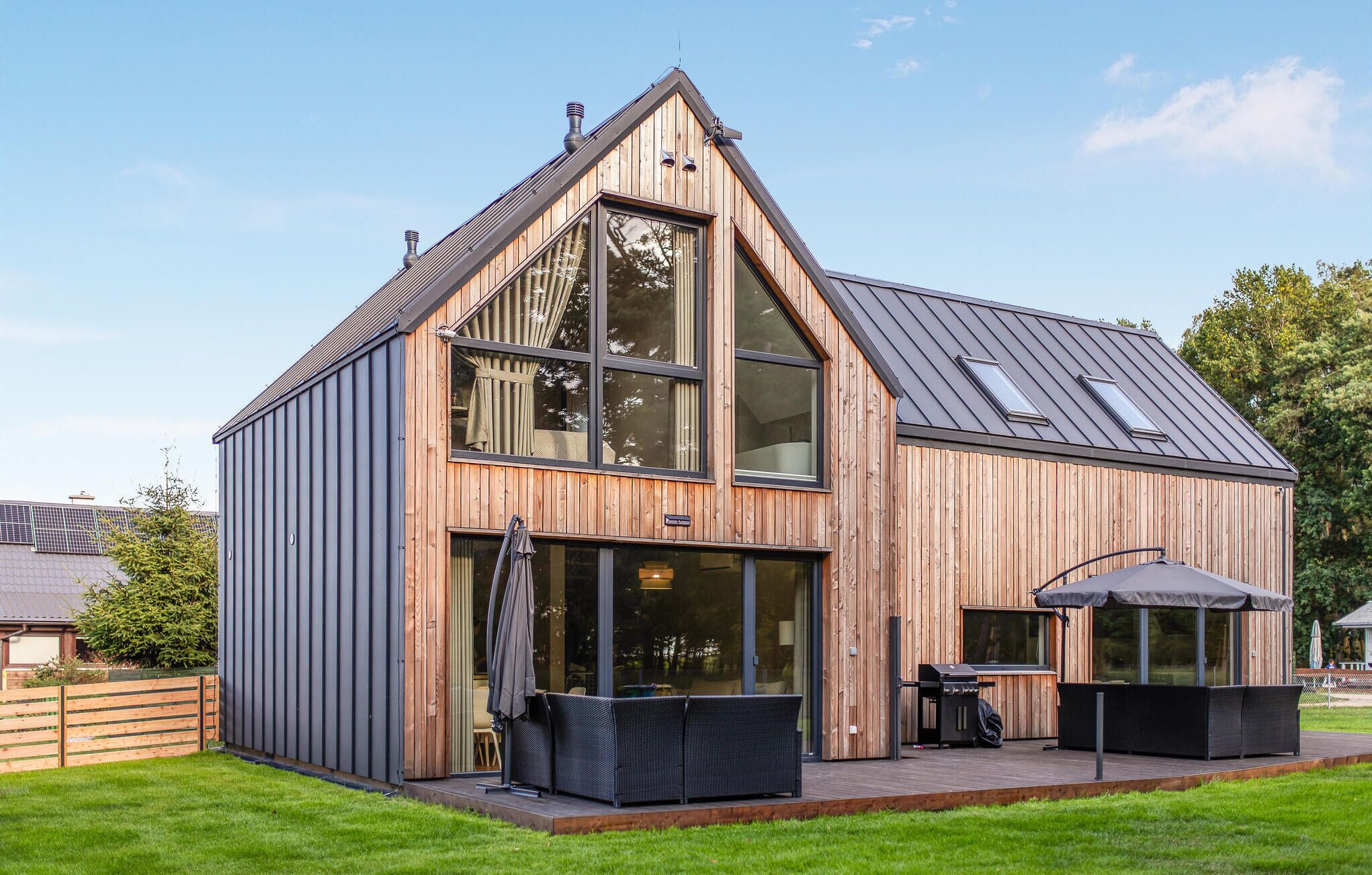 A modern wooden house with a sloped roof, large windows, and an outdoor patio area with furniture.