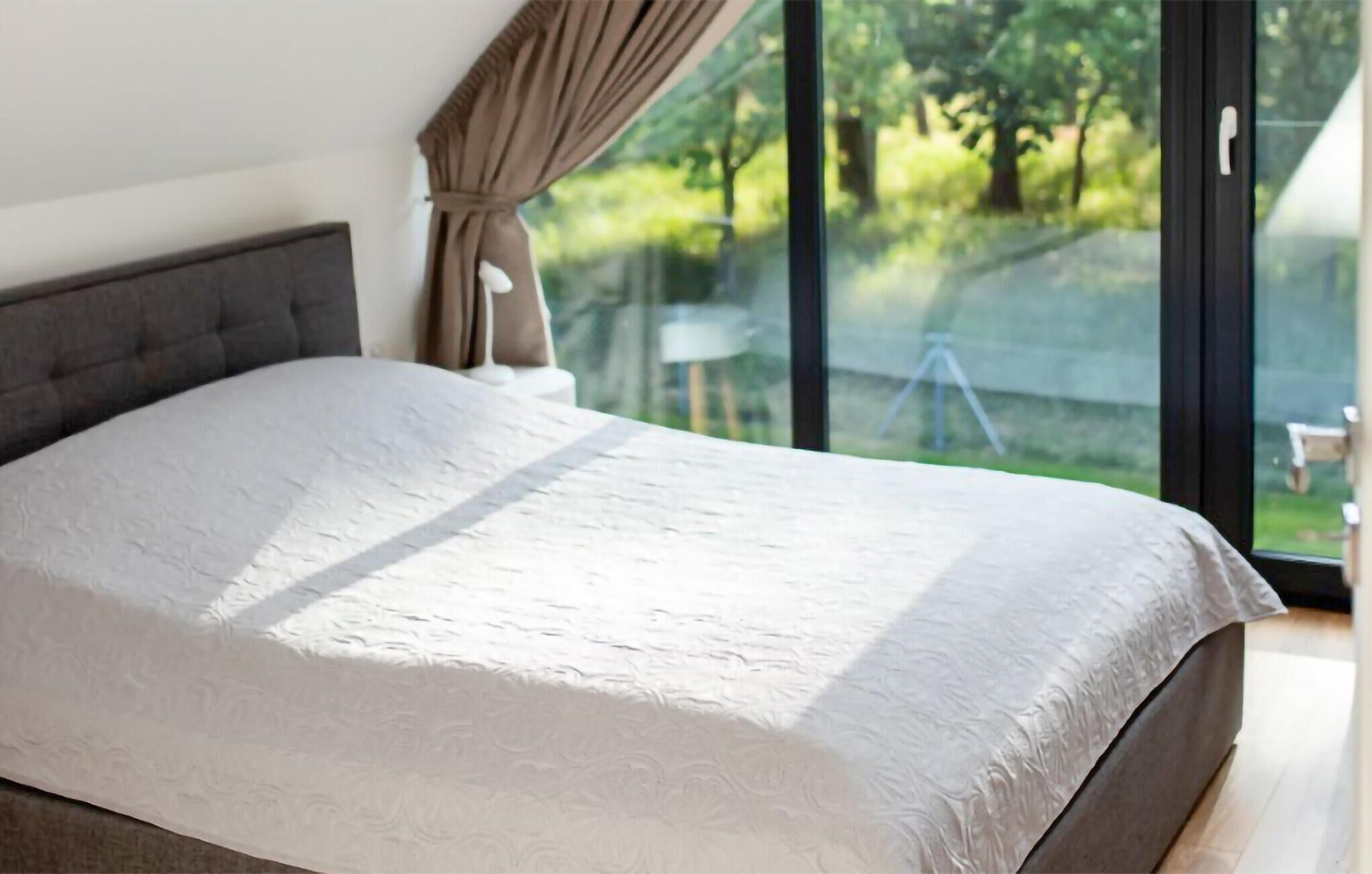 A bedroom with a large bed, a grey headboard, and a view of greenery through large windows.