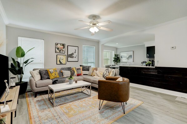 A modern living room with a sofa, coffee table, and a brown armchair. There is a ceiling fan, a TV, and a kitchen area in the background.