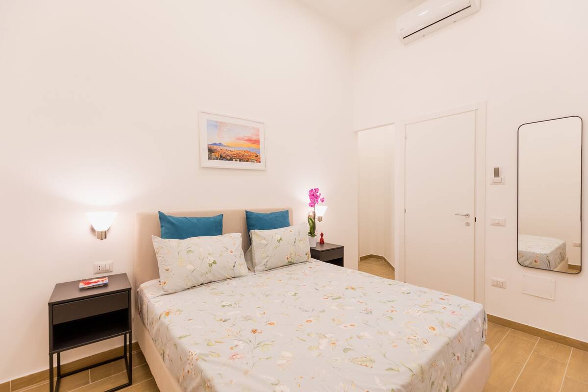 A bedroom with a bed, nightstand, wall-mounted lamp, and a painting.