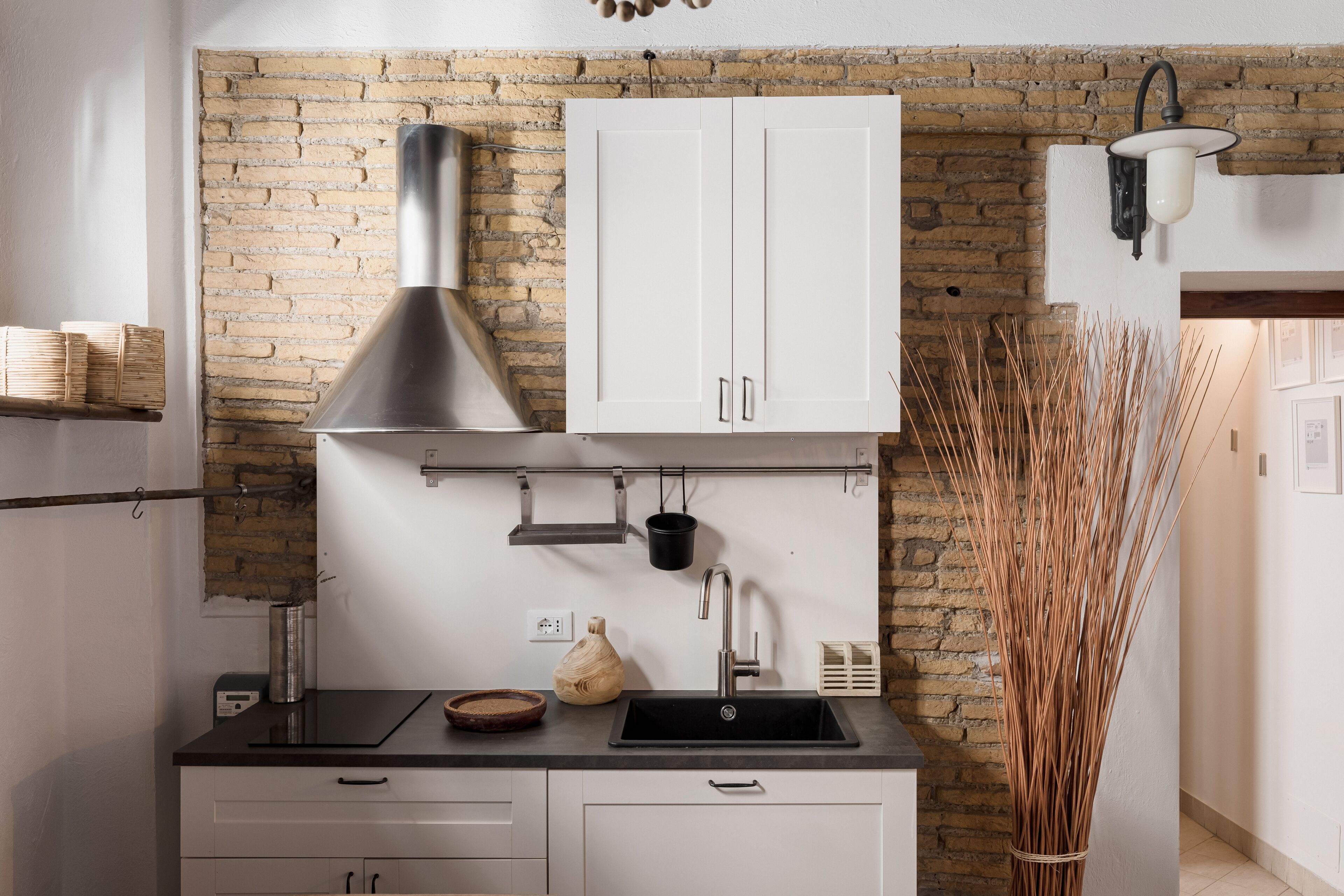 A kitchen with a brick wall, white cabinets, a stainless steel range hood, a black sink, and a hanging pot rack.