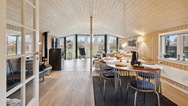 A spacious, well-lit room with a high ceiling and wooden paneling. It features a large dining table with chairs, a fireplace, and large windows providing views of the outdoors. - Lovely home in Strandby with WiFi (Strandby)