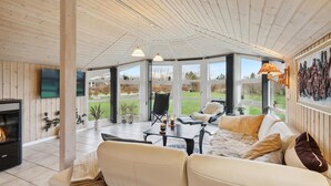 A cozy living room with a fireplace, a sofa, a dining table, and a large window offering a view of the outdoors. - Lovely home in Strandby with WiFi (Strandby)