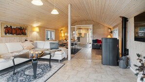 A spacious living area with a sofa, coffee table, and pendant lights. A kitchen is visible in the background, and a fireplace is present. - Lovely home in Strandby with WiFi (Strandby)