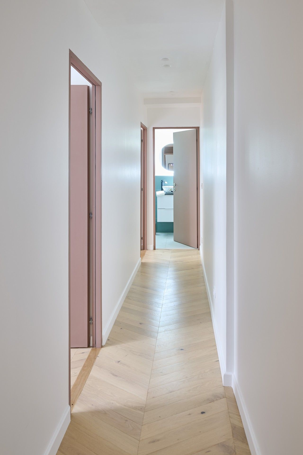 Un pasillo con piso de madera y paredes blancas que conduce a un baño con inodoro y lavamanos.