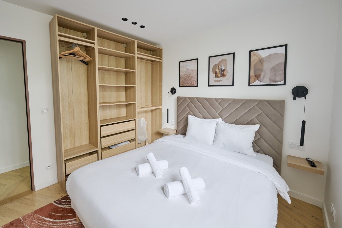 A hotel room with a large bed, a wardrobe with multiple shelves, a bedside table with a lamp, and artwork on the wall.