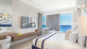 Een moderne hotelkamer met een groot bed, een zithoek met een bank en fauteuils, een eettafel en uitzicht op zee.