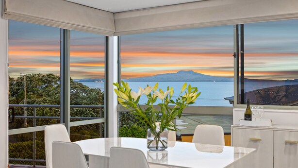 Uma área de jantar moderna com uma mesa grande e cadeiras, um vaso com flores e vista para o pôr do sol sobre o oceano.