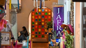 Vista del interior de un restaurante con una entrada colorida, una mujer sentada en una mesa y un letrero de 'JAFRA' al fondo.