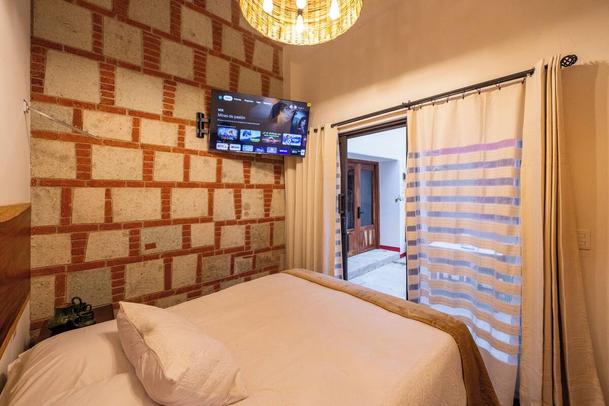 Una habitación de hotel con pared de ladrillo, un televisor de pantalla plana, una cama con almohada blanca y una cortina a rayas.