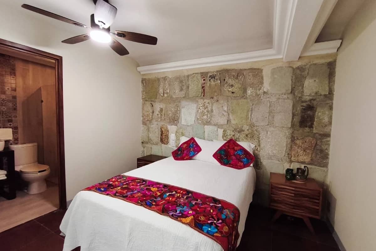 Un dormitorio con cama, ventilador de techo, pared de piedra, mesita de noche de madera y baño con inodoro y lavabo.