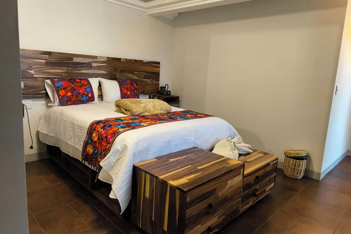 Un dormitorio con cabecera de madera, una cama con edredón floral colorido, una almohada blanca y una mesita de noche de madera con una lámpara.