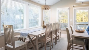 Un espace repas comprenant une table et des chaises, de grandes fenêtres équipées de stores et un luminaire suspendu.