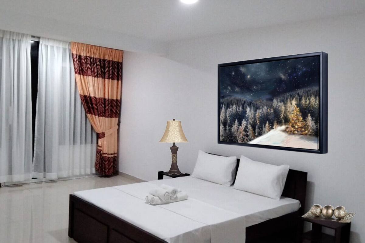 A bedroom with a bed, a nightstand, a lamp, a painting of a snowy forest, and curtains.