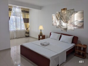 A hotel room with a large bed, a nightstand with a lamp, a wall-mounted artwork, and a window with curtains. - MPM Apartment -Manawickrama Building  (Kandy)