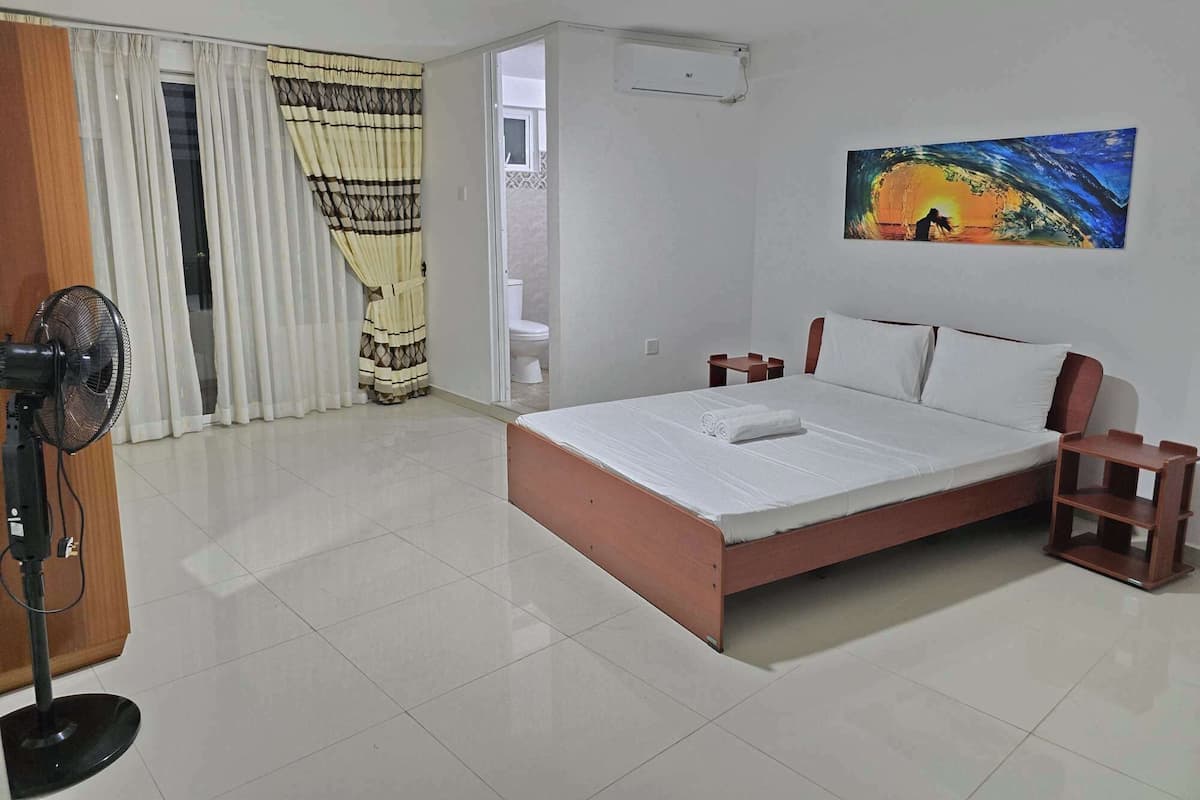A bedroom with a bed, a fan, a painting, and a bathroom visible through an open door.