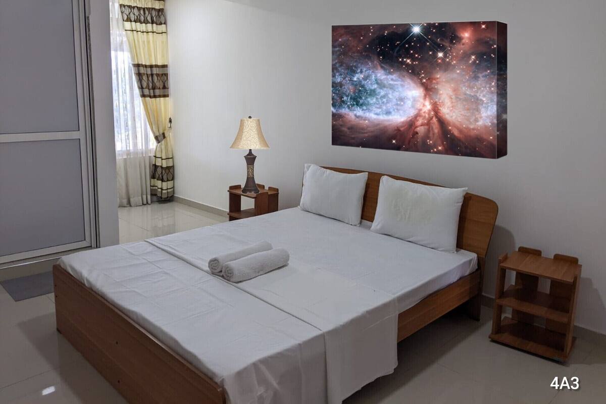 A hotel room with a large bed, a nightstand with a lamp, a wooden nightstand, and a wall-mounted painting of a galaxy.