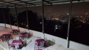 Ein Essbereich im Freien mit Tischen und Stühlen, überdacht und mit Blick auf eine belebte Stadtlandschaft bei Nacht.