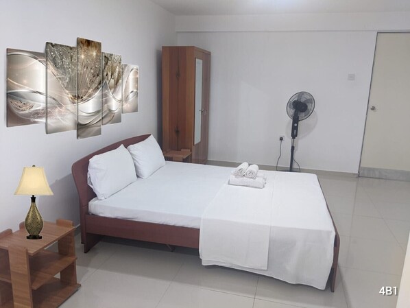 Habitación de hotel ordenada con una cama, una mesita de noche de madera con lámpara, un armario y un ventilador en la pared.