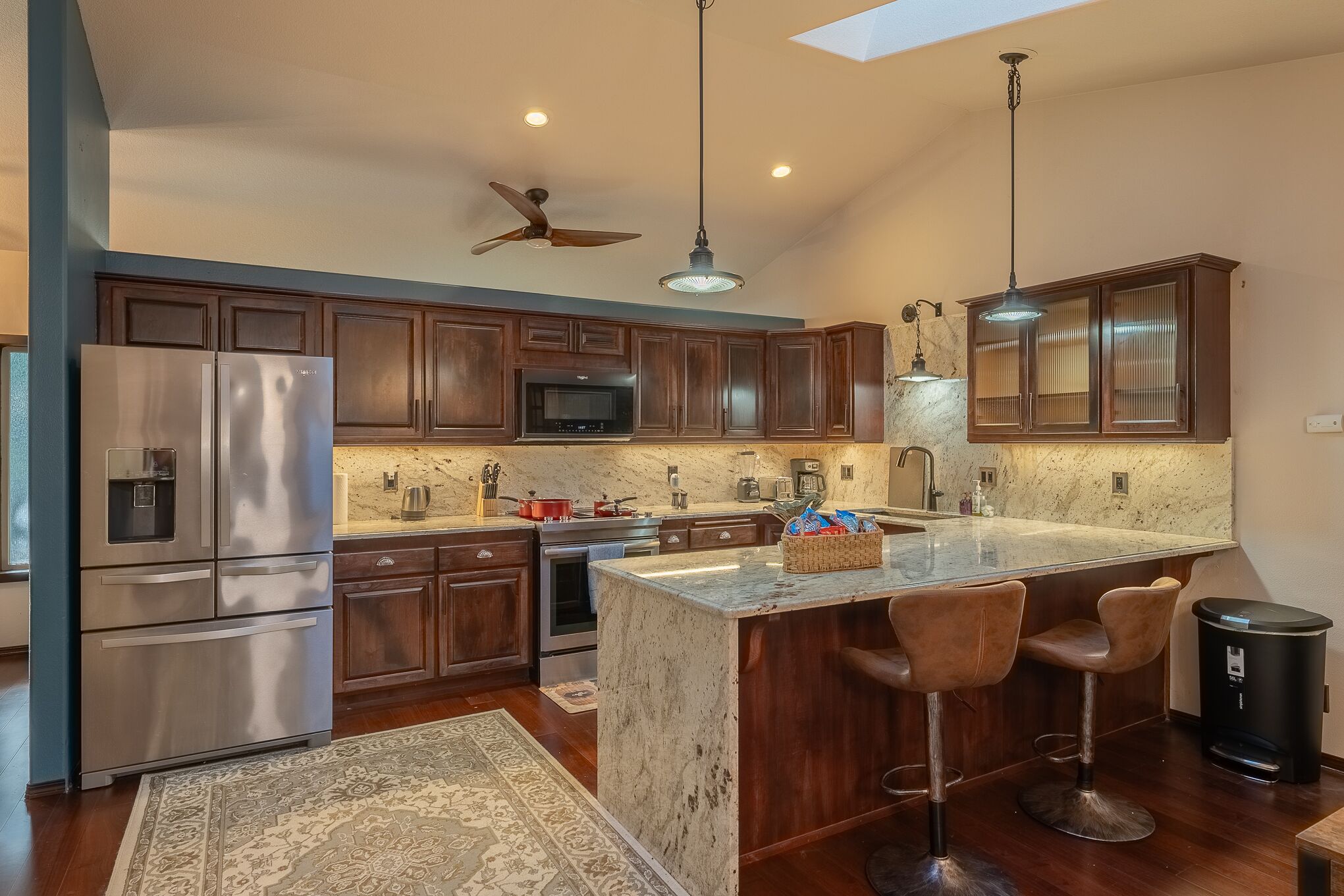 A modern kitchen with dark wooden cabinets, stainless steel appliances, a large island with bar stools, and pendant lighting.