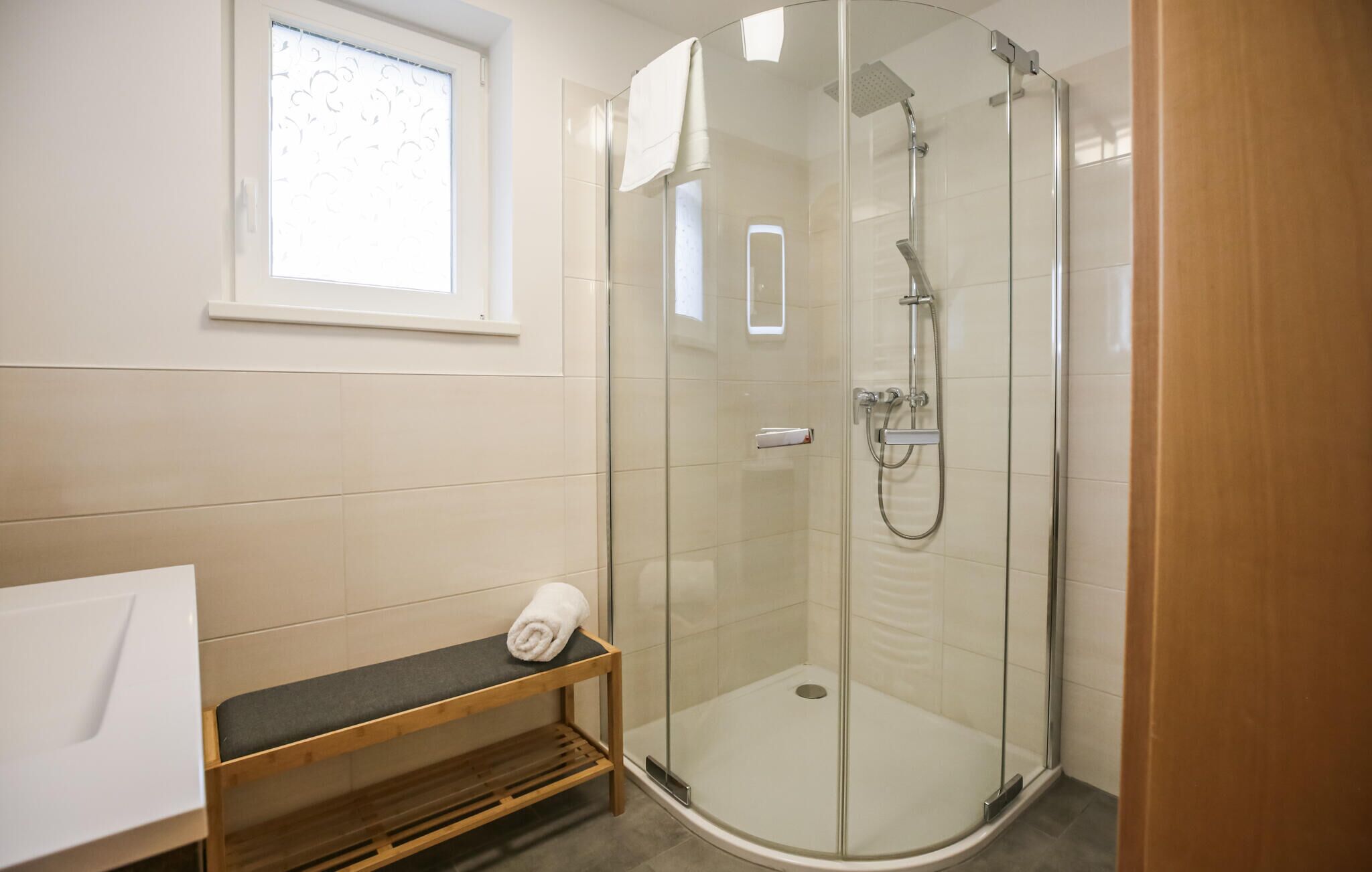 Une salle de bain moderne avec une douche à parois de verre, un banc en bois et un lavabo blanc.