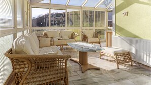 A sunlit room with wicker furniture, a glass-top table, and a view of a mountainous landscape through large windows.